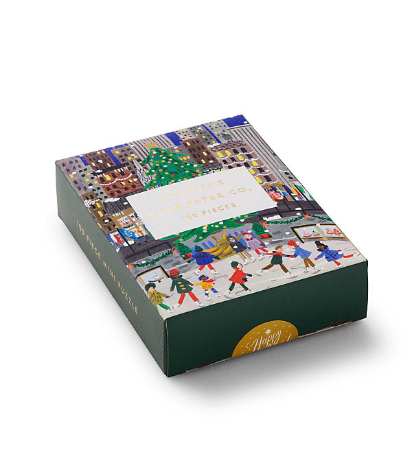 A colorful puzzle box featuring a festive winter city scene with skating figures in the foreground and a decorated tree and buildings in the background, set against a plain white backdrop.