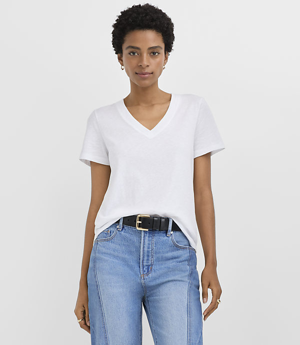 A woman wearing a white V-neck t-shirt and blue jeans with a black belt stands against a plain white background.