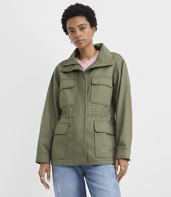 A woman in a green jacket, plaid shirt, denim skirt, and tan boots stands against a plain white background.