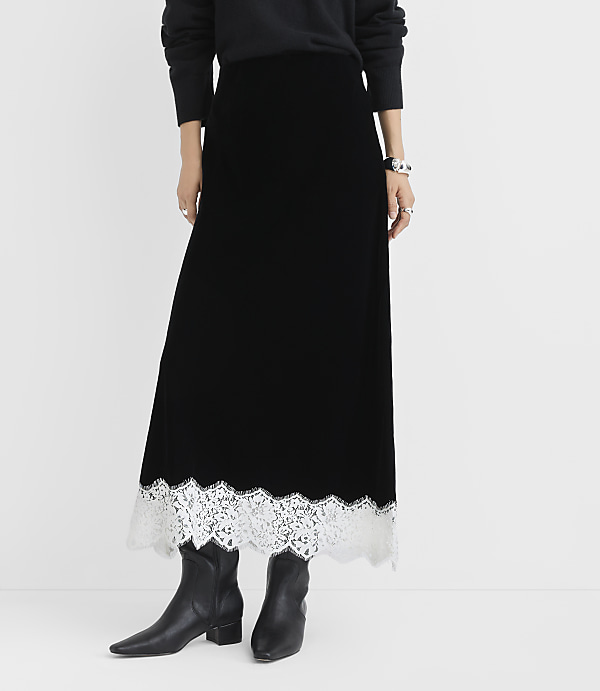 A person wearing a black velvet maxi skirt with a white lace hem and black boots stands against a plain white background.