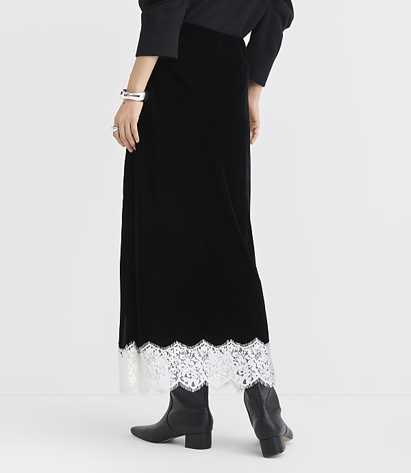 A person wearing a long black velvet skirt with white lace trim and black boots stands against a plain white background.