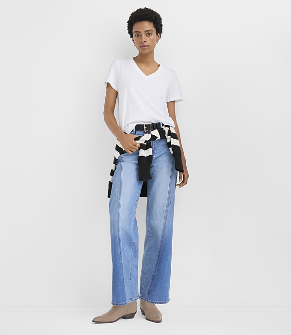 A woman wearing a white v-neck t-shirt, wide-leg blue jeans, and tan boots stands against a plain white background.