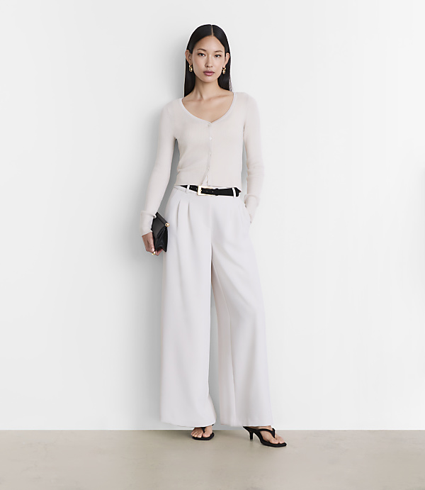 A woman in a cream cardigan and white wide-leg pants stands against a plain white background.