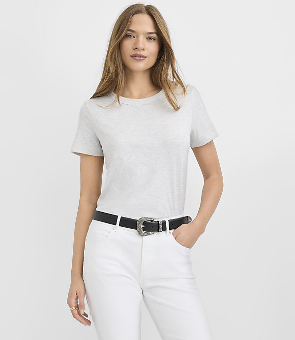A woman wearing a grey t-shirt, white pants, and a black belt stands against a plain white background.