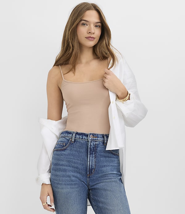 A woman wearing a tan camisole, blue jeans, and an open white shirt stands against a plain white background.