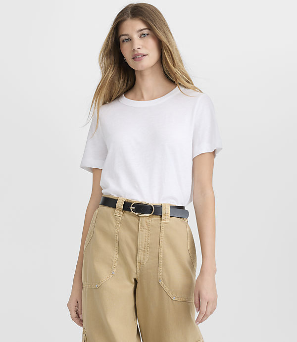 A woman wearing a white t-shirt and khaki pants with a black belt stands against a plain white background.