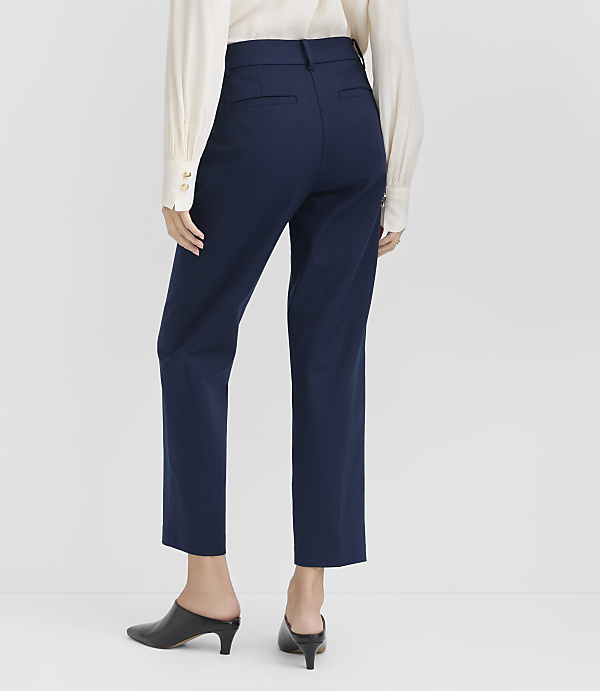 A person wearing a white blouse, navy blue trousers, and black mules stands against a plain light gray background.