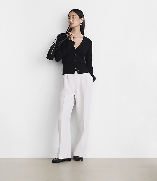 A woman wearing a black cardigan and white wide-leg trousers stands against a plain white wall.