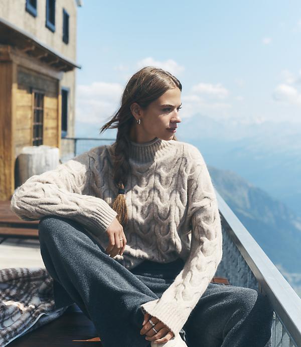 A woman in a beige cable-knit sweater and dark trousers sits on a balcony overlooking a vast mountain landscape.