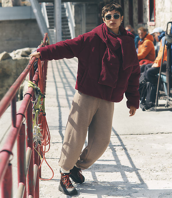 A person wearing a burgundy ribbed sweater and tan trousers leans against a red railing on a sunny concrete walkway with other people sitting in the background.