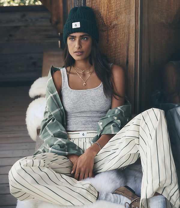 A woman wearing a green beanie, grey tank top, and striped pants sits on a white faux-fur rug against a rustic wooden wall.