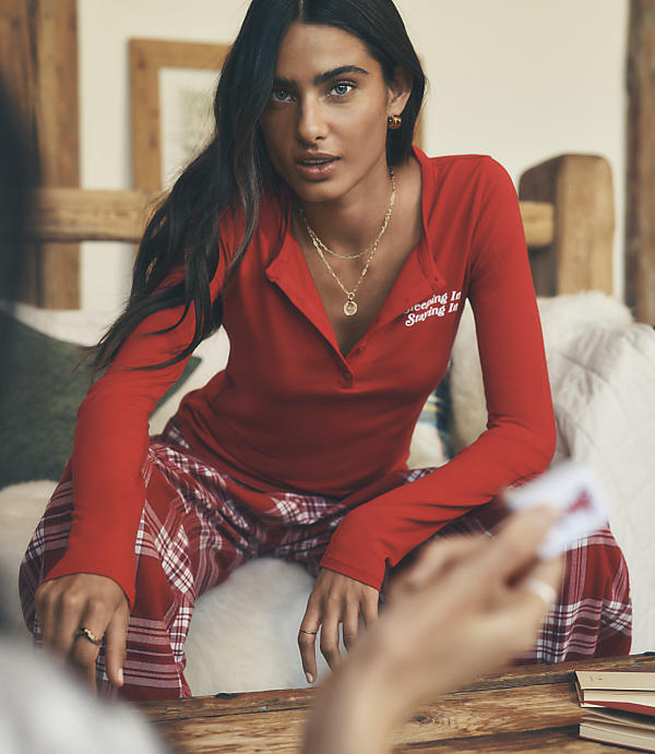 A woman wearing a red long-sleeved shirt and red plaid pajama pants sits on a white fluffy surface in a rustic room with wooden accents.