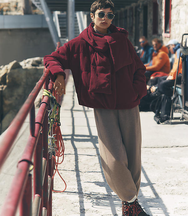 A woman in a bulky burgundy jacket and tan pants leans against a red railing in the foreground, with a concrete walkway and seated people in the background.