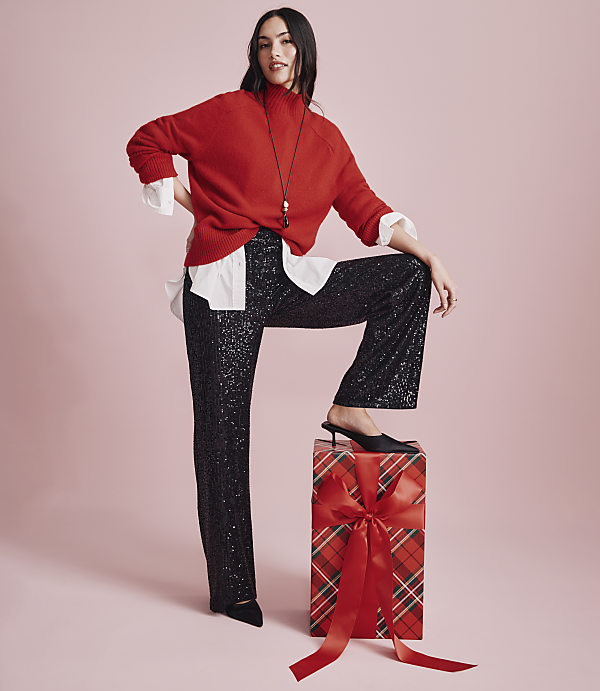 A woman wearing a red sweater and sequined black pants stands with one foot on a large wrapped gift against a solid pink background.