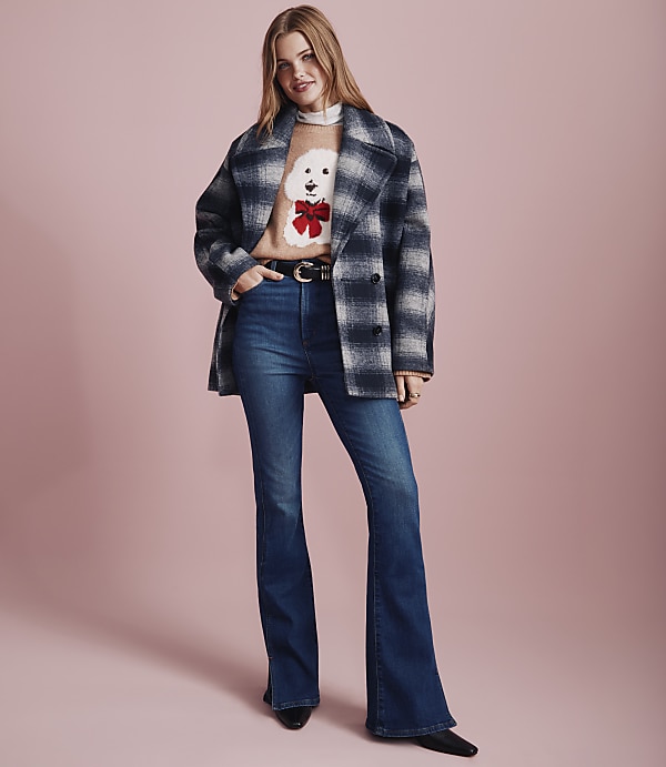 A woman wearing a dog-patterned sweater, a plaid coat, and flared jeans stands against a solid pink background.