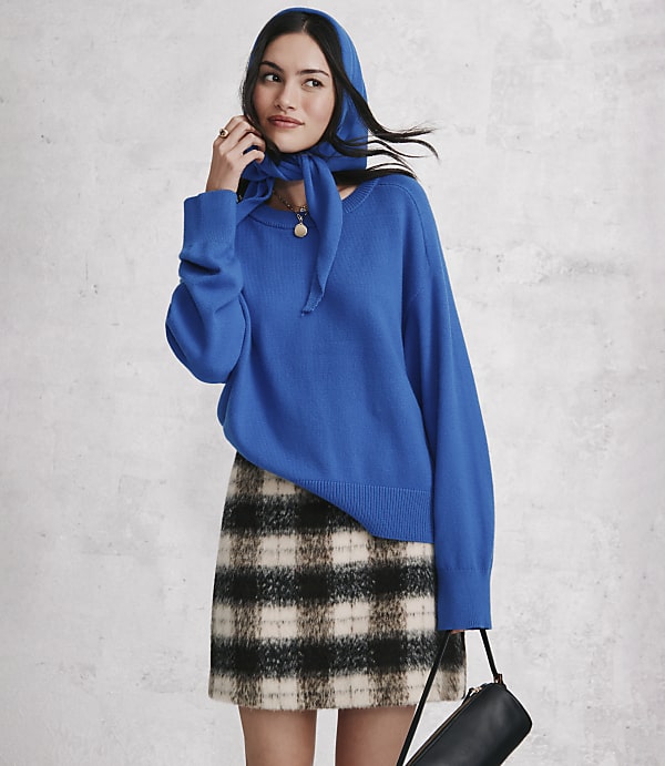 A woman wearing a bright blue sweater, a matching headscarf, and a plaid mini skirt stands against a textured grey wall.