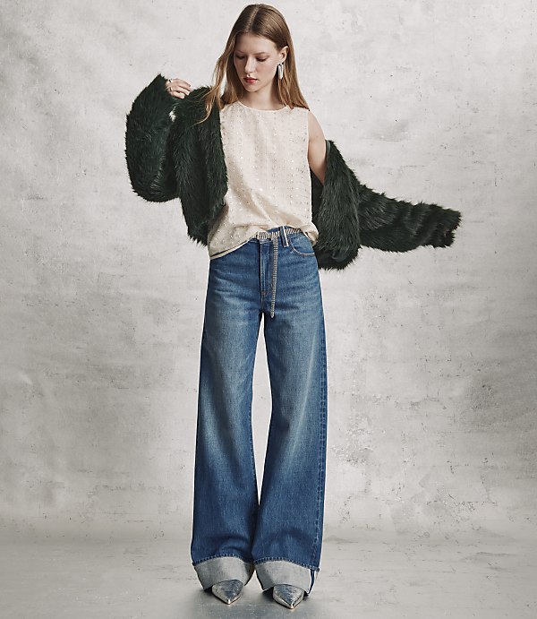 A woman wearing a dark green faux fur coat, a cream top, and wide-leg blue jeans stands against a textured grey concrete wall.