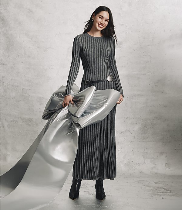A smiling woman in a long, ribbed grey dress holds a large piece of shimmering silver fabric against a plain, textured grey concrete wall.