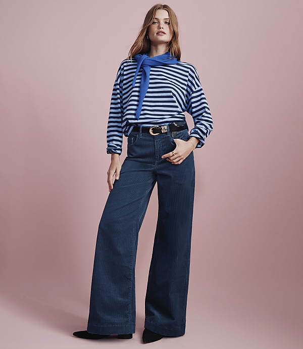 A woman wearing a blue and white striped shirt, a blue neck scarf, and wide-leg blue trousers stands against a solid pink background.