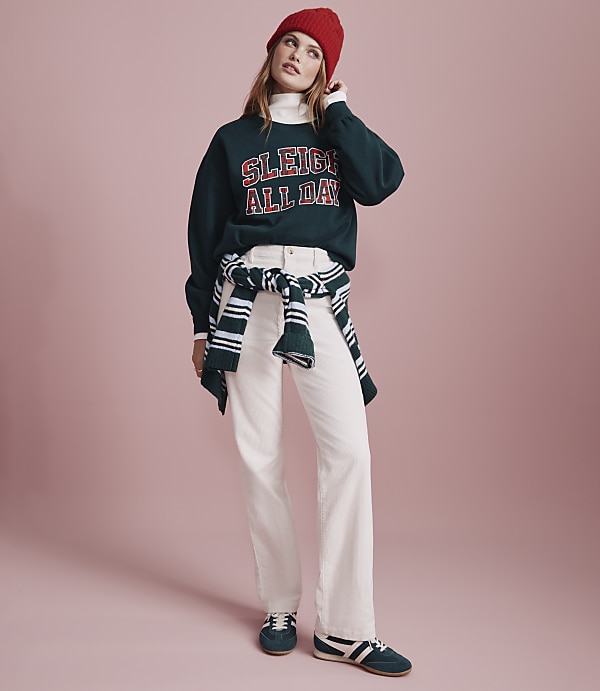 A woman wearing a red beanie, a dark green "Sleigh All Day" sweatshirt, and white pants stands against a solid pink background.