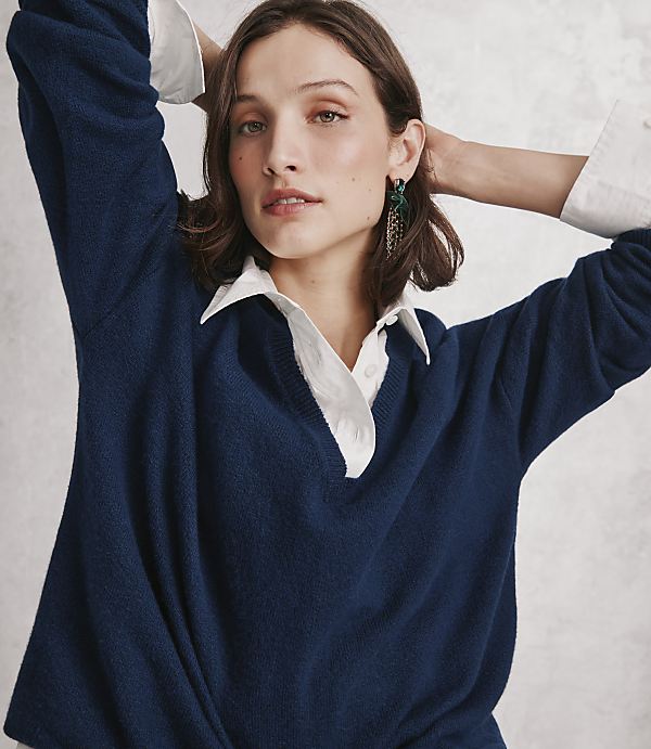 A woman wearing a navy blue sweater over a white collared shirt poses with her arms raised against a plain, light-grey textured wall.