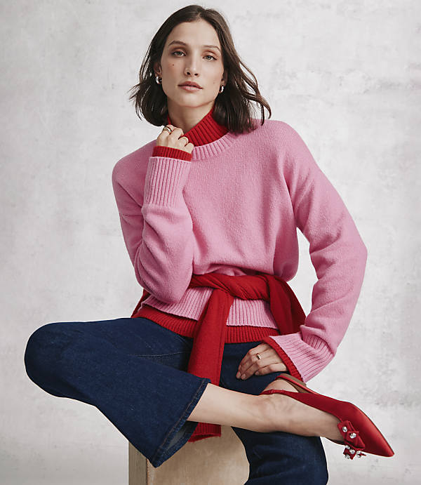 A woman wearing a pink sweater, red turtleneck, and blue jeans sits against a neutral, textured grey wall.