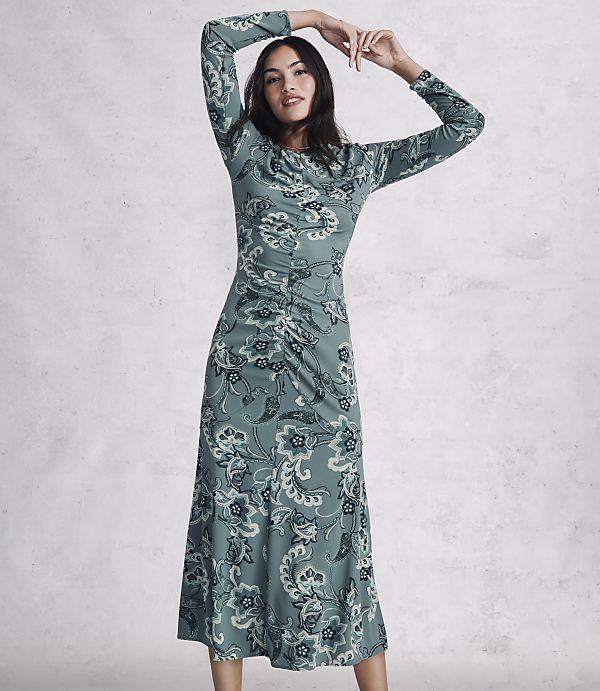 A woman in a long-sleeved, floral-patterned green dress poses with her arms raised against a textured grey wall.