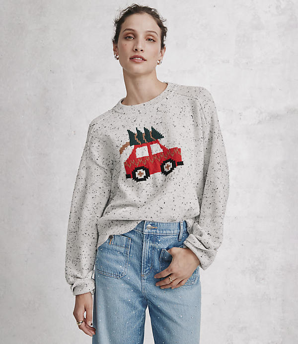 A woman wearing a speckled grey sweater with a red car and Christmas tree design and blue jeans stands against a plain, light grey concrete wall.