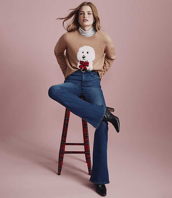 A woman wearing a dog-patterned sweater and blue jeans sits on a red plaid stool against a solid pink background.