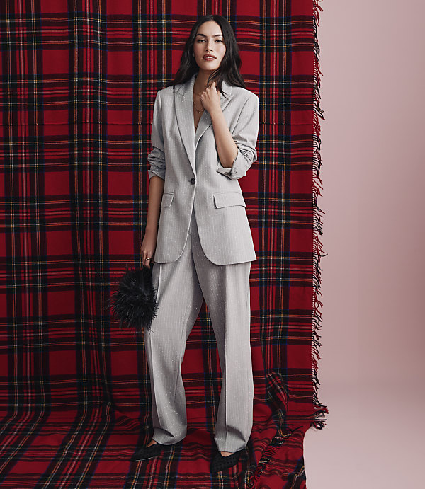 A woman in a light grey pant suit stands in the foreground against a large red and black plaid backdrop.