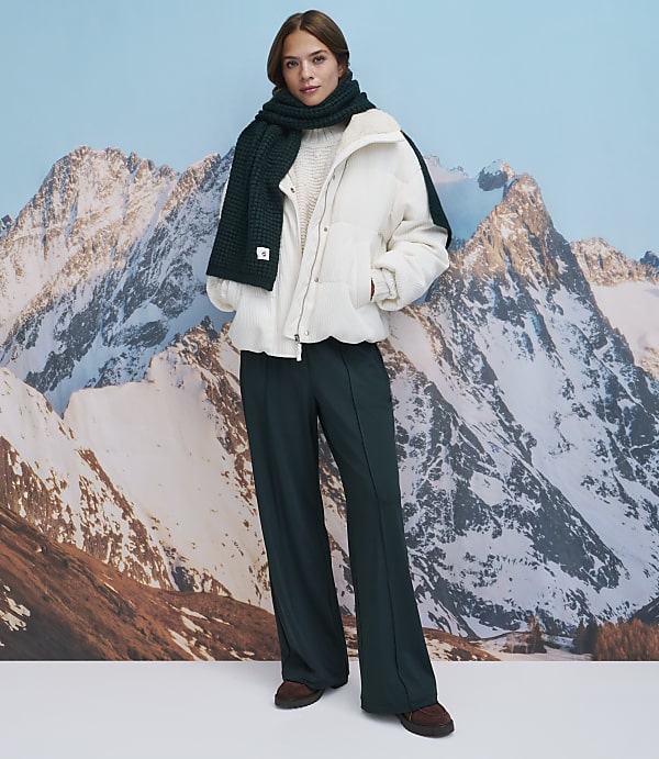 A woman wearing a white puffer jacket, dark green wide-leg trousers, and a matching scarf stands before a backdrop of snow-capped mountains.