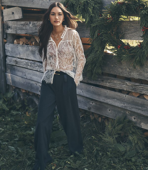A woman in a white lace blouse and black trousers stands in the foreground against a background of a rustic wooden fence adorned with evergreen branches.