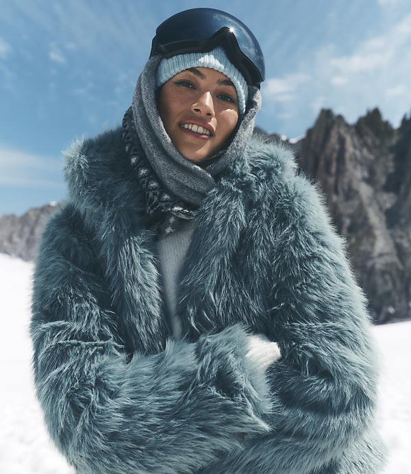 A woman wearing a blue faux-fur coat, beanie, and ski goggles stands against a backdrop of snowy mountains and a clear blue sky.