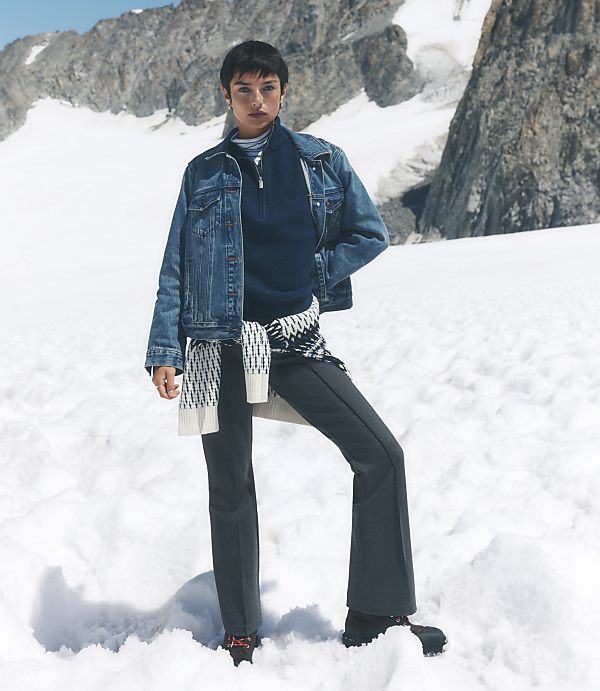 A woman wearing a denim jacket and dark trousers stands in a snowy foreground against a backdrop of rugged, snow-capped mountains.