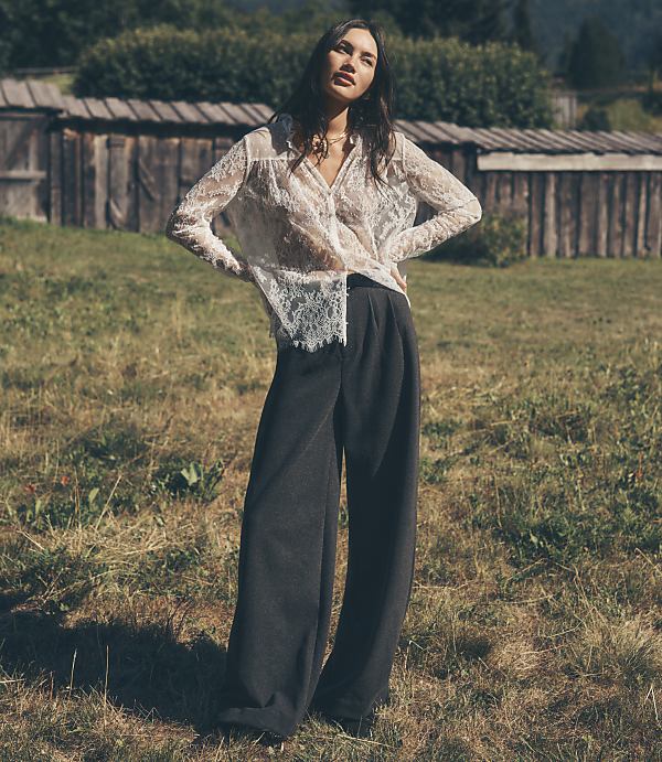A woman wearing a white lace blouse and wide-leg dark trousers stands in a grassy field in front of a rustic wooden fence.
