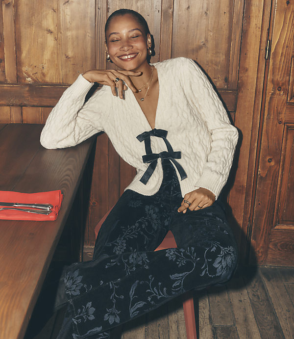 A smiling woman wearing a white cable-knit cardigan and dark floral trousers sits at a wooden table against a rustic wood-paneled wall.