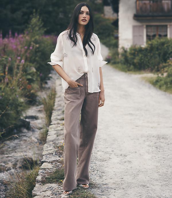 A woman in a white embellished blouse and brown wide-leg trousers stands on a gravel path surrounded by lush greenery and a rustic building.