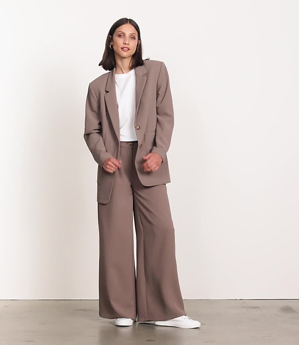 A woman in a black blazer and wide-leg pants stands against a plain white background.