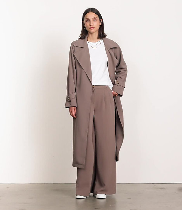 A woman in a plum trench coat and matching pants stands with a dark handbag against a plain white background.
