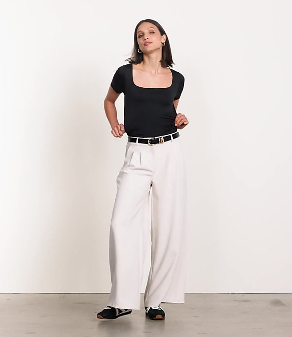 A woman in a cream-colored tank top, cardigan, and wide-leg pants stands against a plain white background.